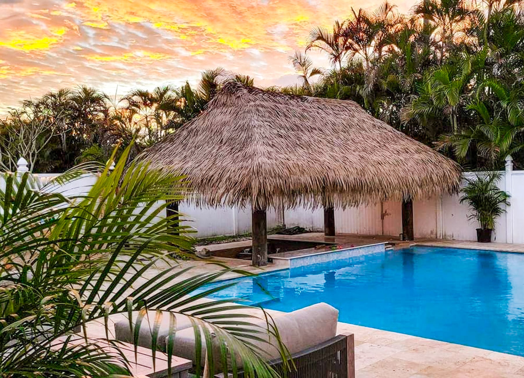 Synthetic thatch palapa next to a pool under a colorful sunset.