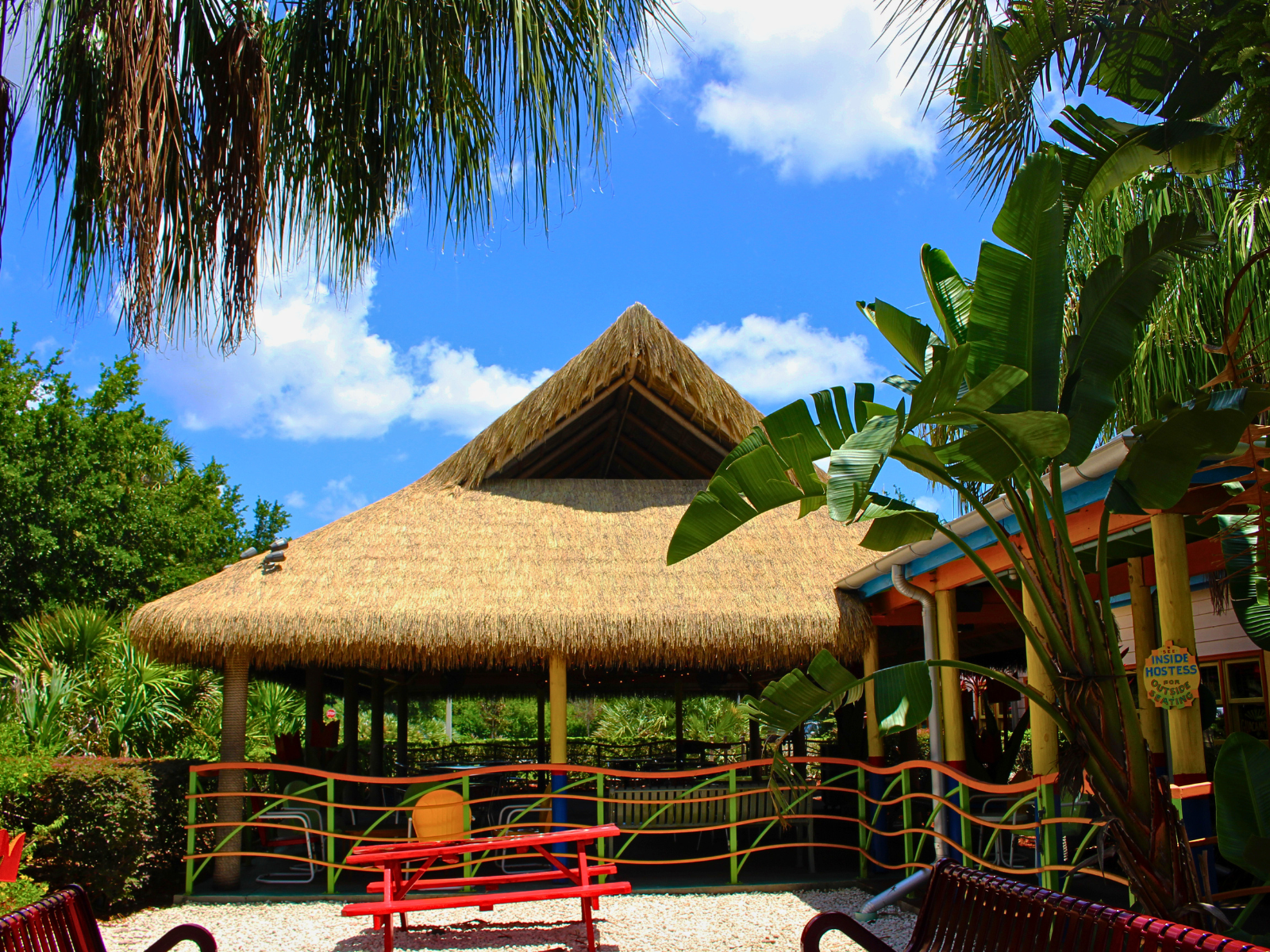 Large Endureed Premium Bali thatch-roof structure at Chuy's restaurant, framed by palm trees and tropical banana plants under a bright blue sky.