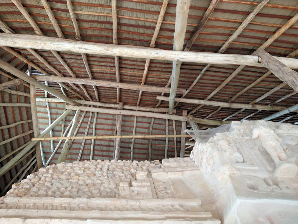 Interior underside view of Endureed Performance Weathered Palm synthetic thatch installation at Ek Balam Mexico &mdash; wood beam and purlin framing structure overhead protecting Mayan carved stone reliefs and stucco sculptures