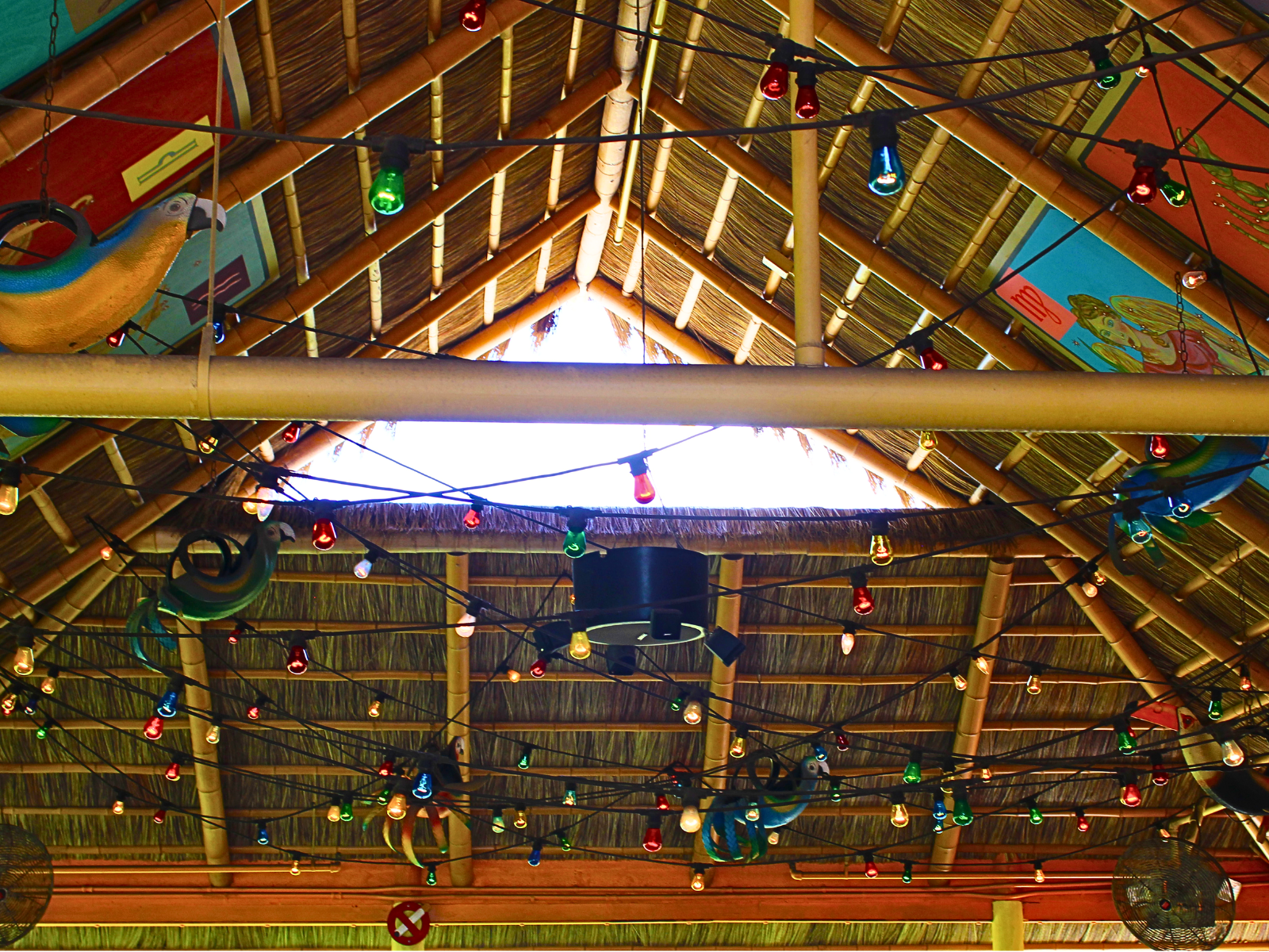 Upward view of an Endureed Premium Bali thatch roof interior at Chuy's restaurant, showing bamboo rafters, layered thatch panels, colorful string lights, and a ceiling fan