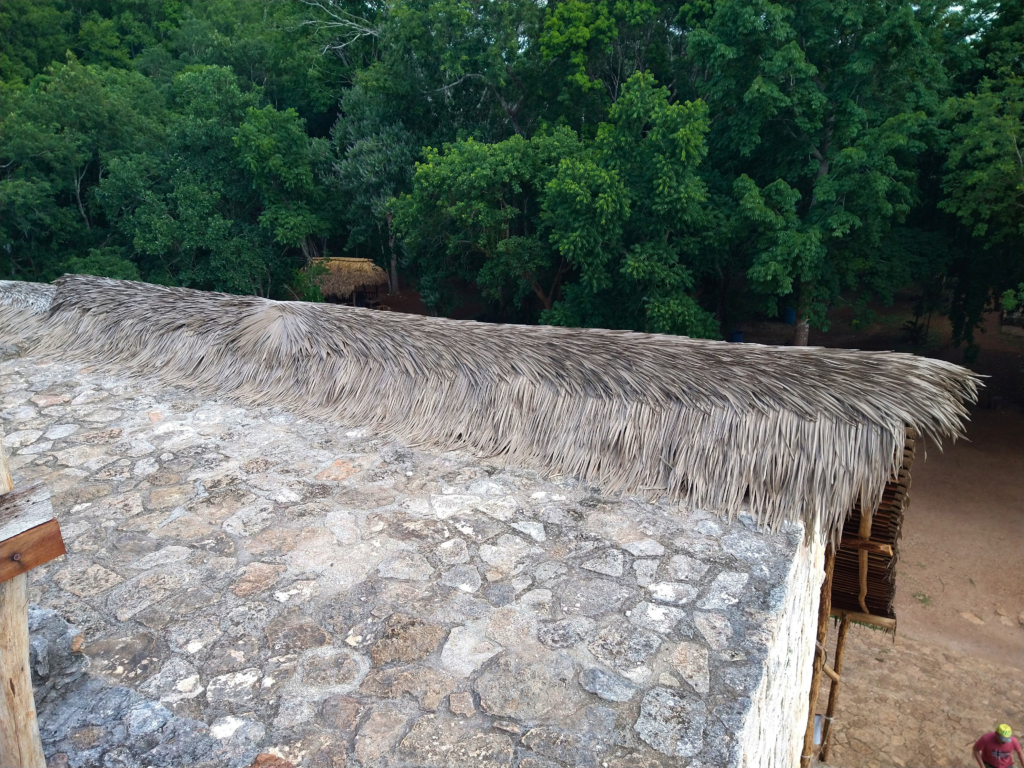 Aerial close-up of Endureed Performance Weathered Palm engineered thatch ridgeline and eave over Ek Balam ancient stone masonry &mdash; layered palm frond panels and bamboo framing with Yucatan jungle canopy in background