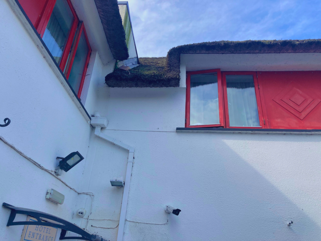 Upward angle of the Thatch Bar's thatched roofline from the covered car park entrance, showing natural thatch fronds along the eave edge against a bright blue sky and red window surrounds.