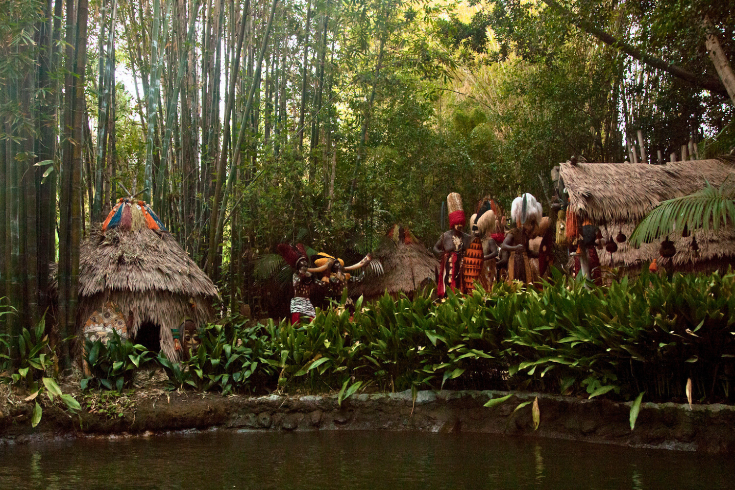 Wide scene view of Endureed Premium Dominica synthetic thatch structures at Disneyland Adventureland &mdash; multiple thatched huts with lush bamboo forest and animated figures alongside water