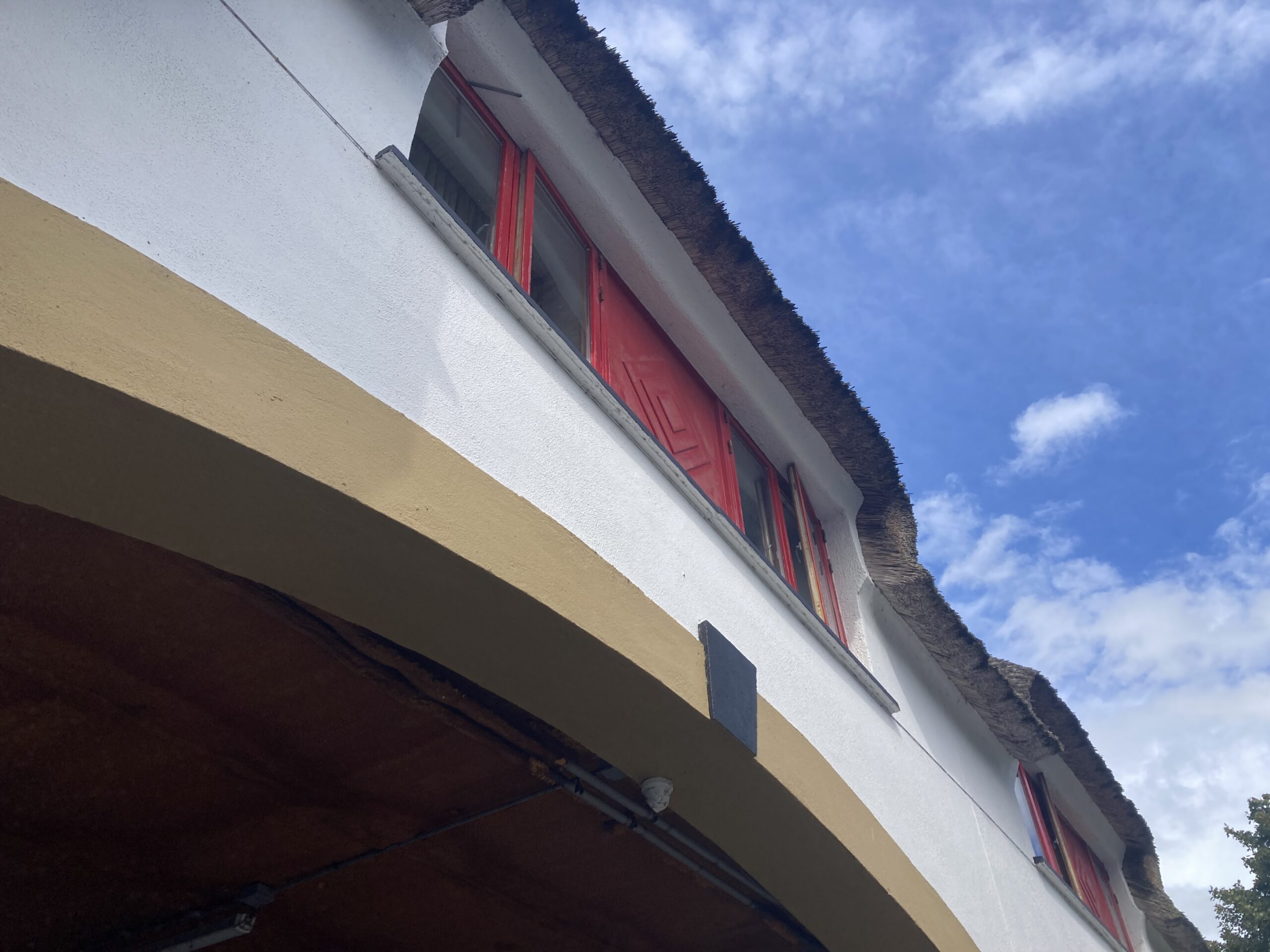 Upward angle of the Marriman Hotel's thatched roofline from the covered car park entrance, showing natural thatch fronds along the eave edge against a bright blue sky and red window surrounds.