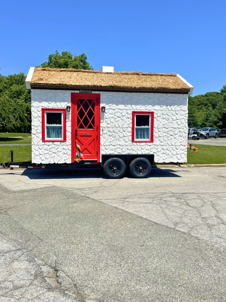 The Thatch Pub mobile Irish bar with Endureed Premium Somerset synthetic thatch roofing &mdash; traveling thatched pub on trailer with white stone exterior red door and windows parked in sunny parking lot Staten Island New York