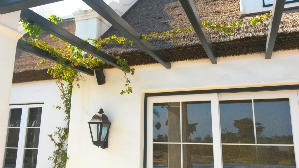 Close-up of Endureed Premium Kilimanjaro engineered thatch eave and pergola framework at Dean's Grange Malibu California &mdash; synthetic thatch roofline with dark timber beams and climbing vine on white stucco exterior