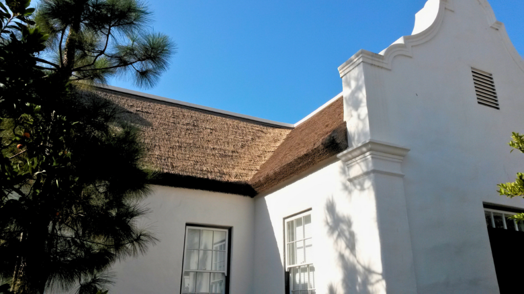 Endureed Premium Kilimanjaro engineered thatch roof on Cape Dutch gable elevation at Dean's Grange Malibu California &mdash; synthetic thatch covering hip roof with ornate curved white parapet against clear blue sky