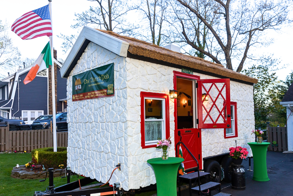 The Thatch Pub traveling Irish bar with Endureed Premium Somerset engineered thatch roofing at evening event &mdash; mobile thatched pub on trailer with American and Irish flags red door open and party lighting in residential setting