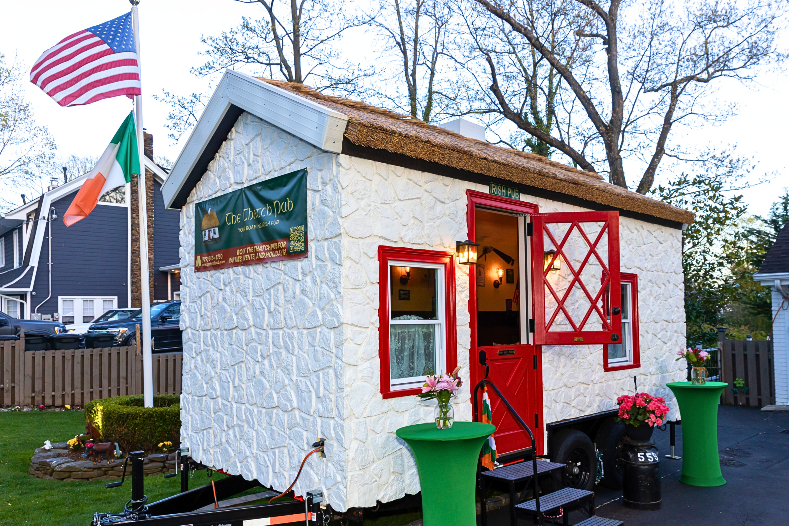 The Thatch Pub traveling Irish bar with Endureed Premium Somerset engineered thatch roofing at evening event &mdash; mobile thatched pub on trailer with American and Irish flags red door open and party lighting in residential setting