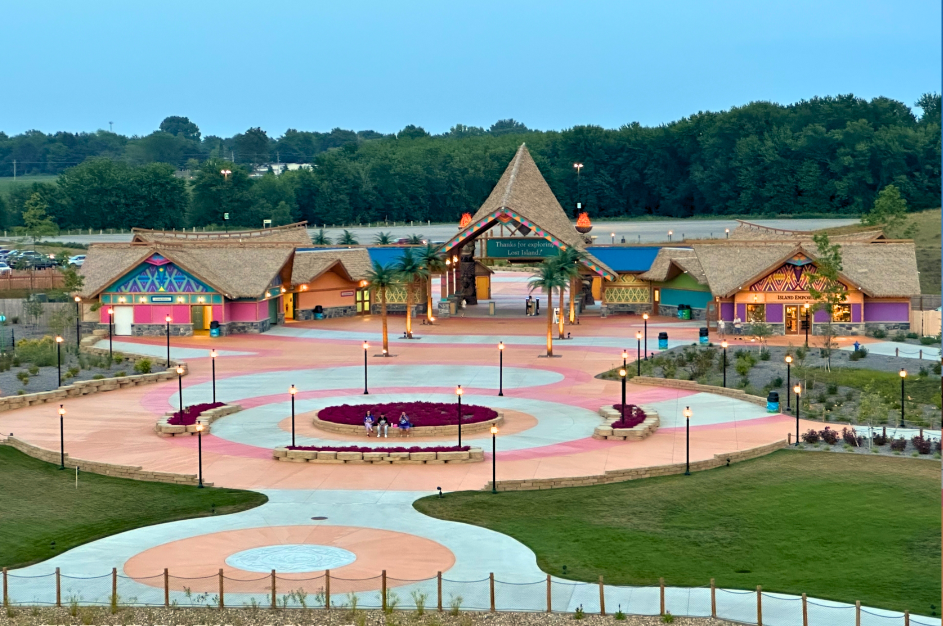 Lost Island Theme Park exit plaza in Waterloo Iowa at dusk featuring Endureed Performance Palm synthetic thatch spanning the full entrance building complex &mdash; engineered thatch roofline illuminated across tribal-themed gateway with colorful murals