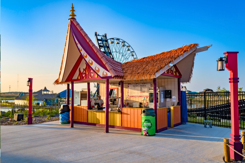 Endureed Performance Palm synthetic thatch on Kotaki Treats food stand at Lost Island Theme Park Waterloo Iowa &mdash; engineered thatch hip roof on open-air snack kiosk with decorative flame gable and Ferris wheel in background