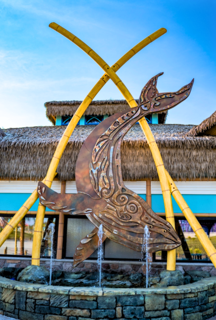 Endureed Performance Palm synthetic thatch roofline at Lost Island Theme Park Waterloo Iowa &mdash; engineered thatch fringe detail visible behind decorative copper whale sculpture fountain with crossed bamboo poles in foreground