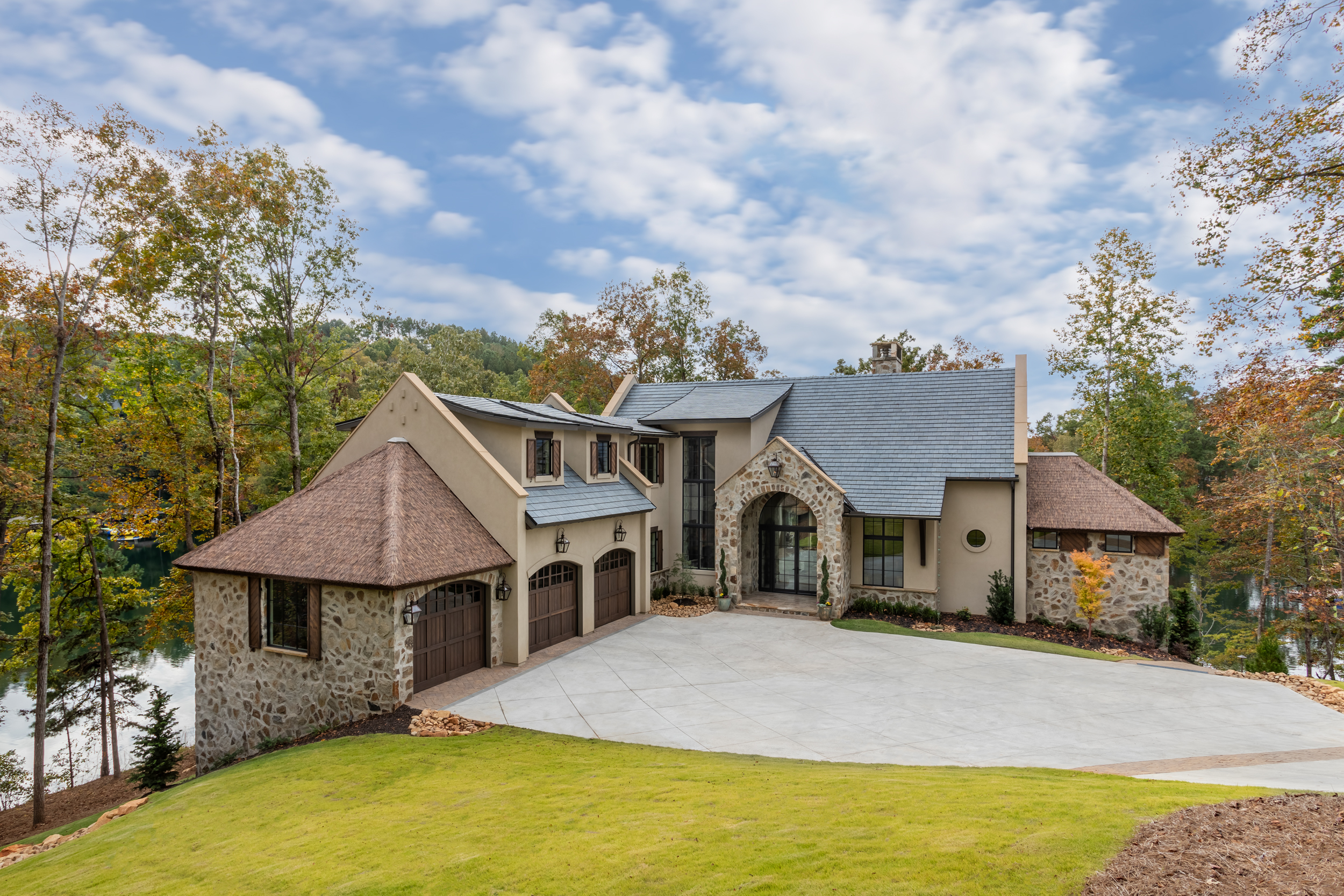The Pendry private residence at Lake Keowee South Carolina featuring Endureed Premium Kilimanjaro synthetic thatch on flanking turret structures &mdash; engineered thatch tower roofs alongside slate main roof on luxury French country lakefront estate with fall foliage. IMKM Architects