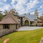 how does synthetic thatch work | Endureed Premium Kilimanjaro synthetic thatch on twin turret roofs at The Pendry Lake Keowee South Carolina &mdash; engineered thatch flanking the stone and stucco luxury lakefront residence with fall color forest backdrop and curved driveway