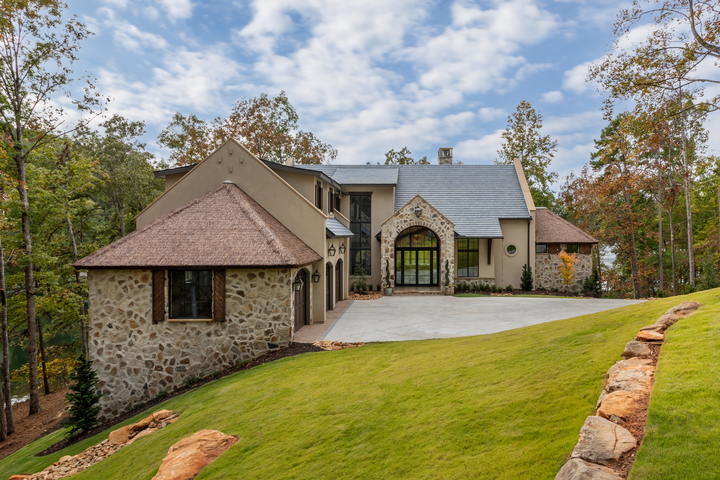 how does synthetic thatch work | Endureed Premium Kilimanjaro synthetic thatch on twin turret roofs at The Pendry Lake Keowee South Carolina &mdash; engineered thatch flanking the stone and stucco luxury lakefront residence with fall color forest backdrop and curved driveway