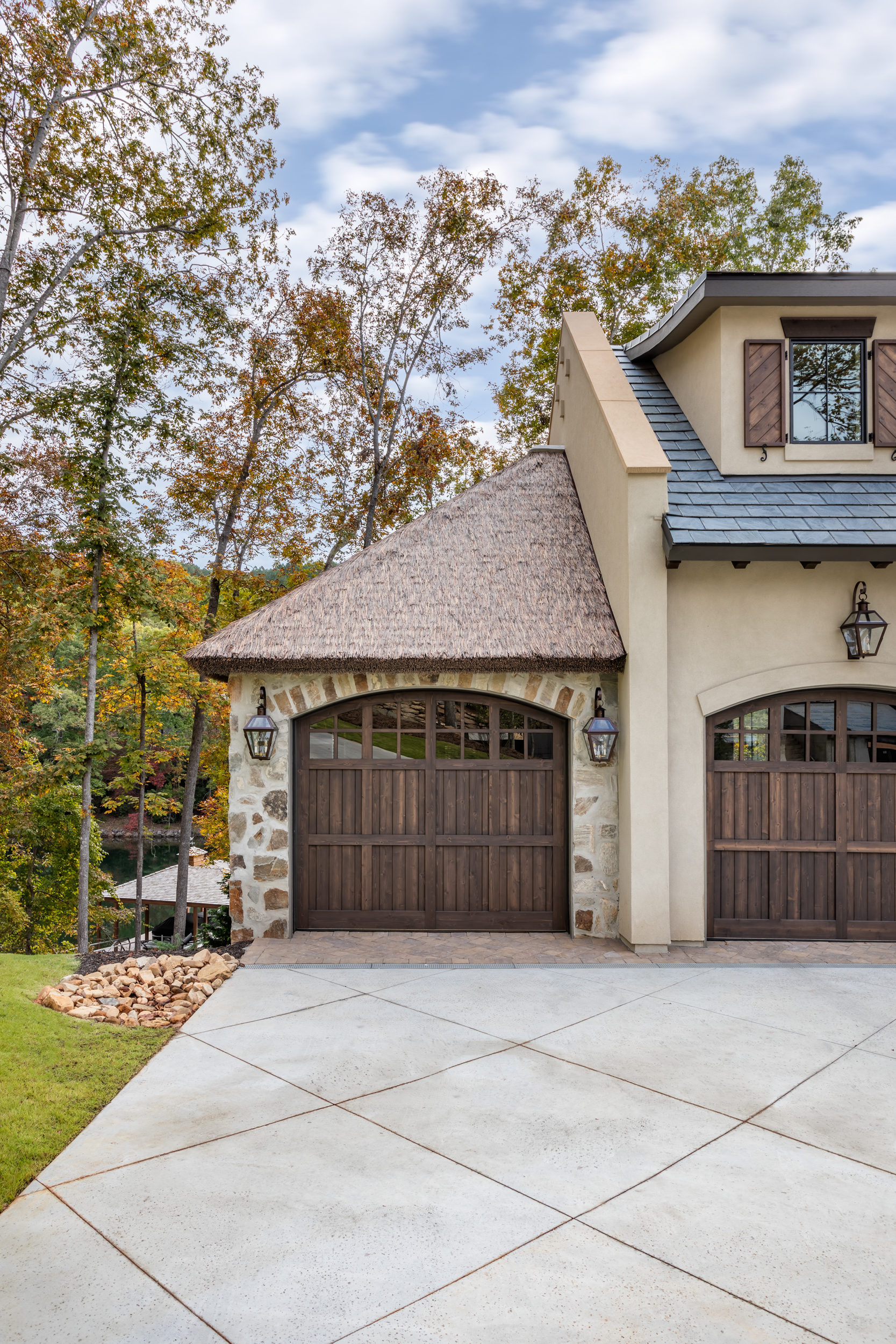 Close-up of Endureed Premium Kilimanjaro synthetic thatch hip roof on garage turret at The Pendry Lake Keowee South Carolina &mdash; engineered thatch conical roof over stone arch and carriage-style garage doors with fall foliage and lake view