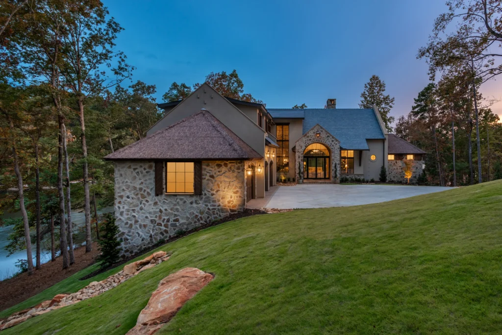 Twilight view of The Pendry luxury residence at Lake Keowee South Carolina &mdash; Endureed Premium Kilimanjaro synthetic thatch turret roofs glowing at dusk flanking illuminated stone and stucco lakefront estate with autumn trees