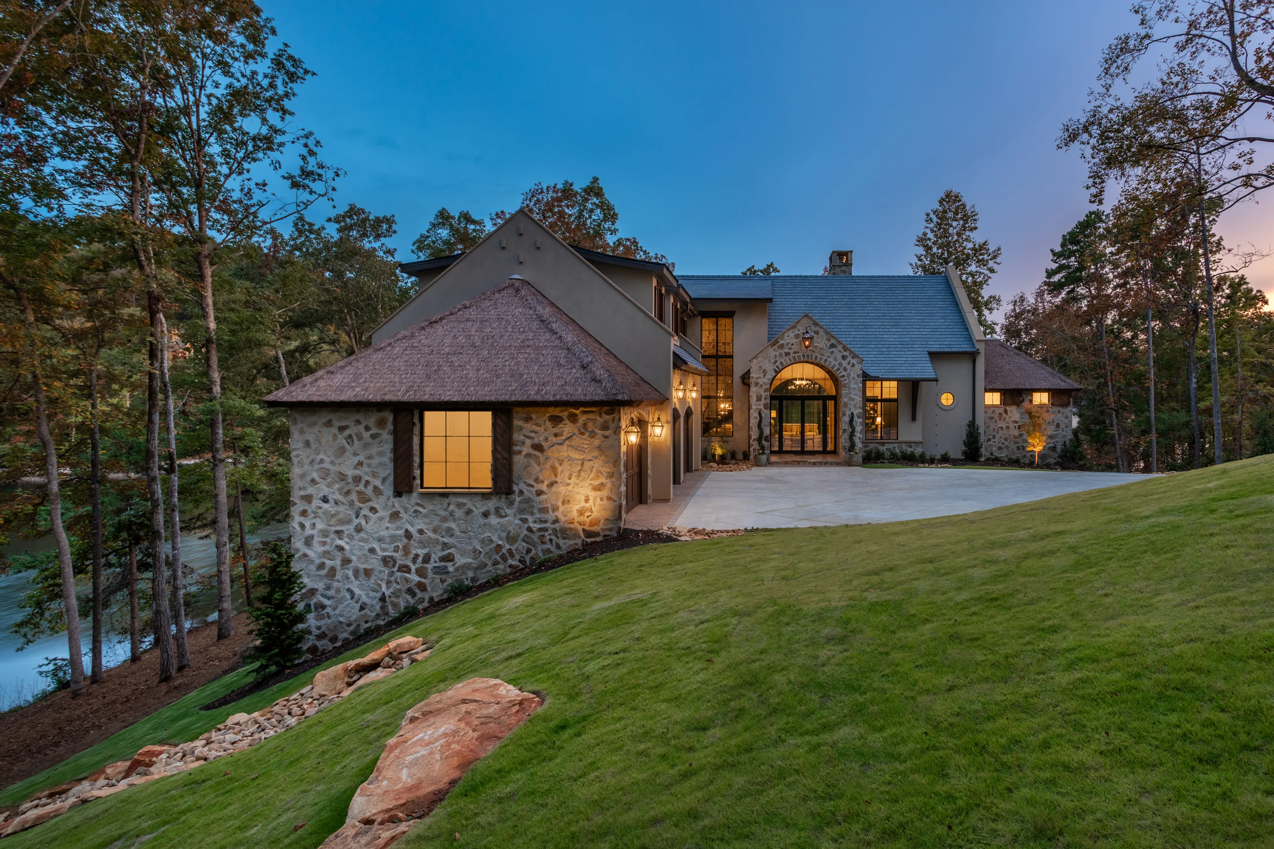 Twilight view of The Pendry luxury residence at Lake Keowee South Carolina — Endureed Premium Kilimanjaro synthetic thatch turret roofs glowing at dusk flanking illuminated stone and stucco lakefront estate with autumn trees