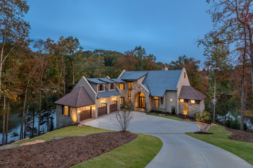 Dusk aerial view of The Pendry lakefront estate at Lake Keowee South Carolina &mdash; Endureed Premium Kilimanjaro engineered thatch on flanking turret structures with illuminated interior windows and autumn forest surrounding the luxury lake house