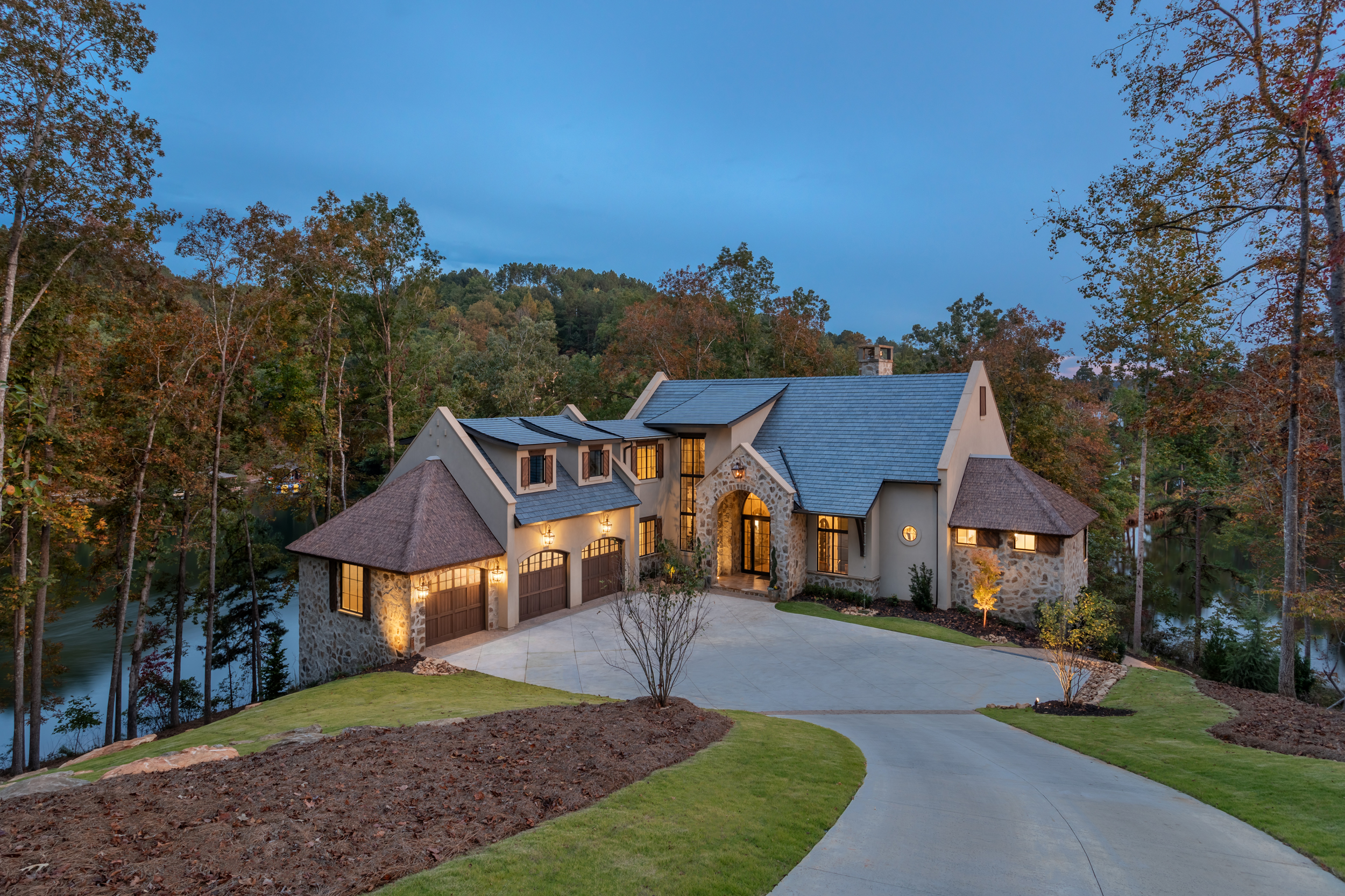 Dusk aerial view of The Pendry lakefront estate at Lake Keowee South Carolina — Endureed Premium Kilimanjaro engineered thatch on flanking turret structures with illuminated interior windows and autumn forest surrounding the luxury lake house