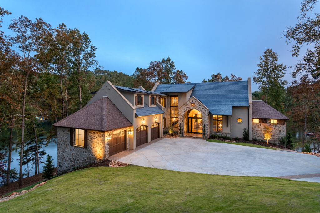 Evening exterior of The Pendry private residence Lake Keowee South Carolina featuring Endureed Premium Kilimanjaro synthetic thatch turret roofs &mdash; engineered thatch on side towers with warm interior lighting and lake visible through autumn trees