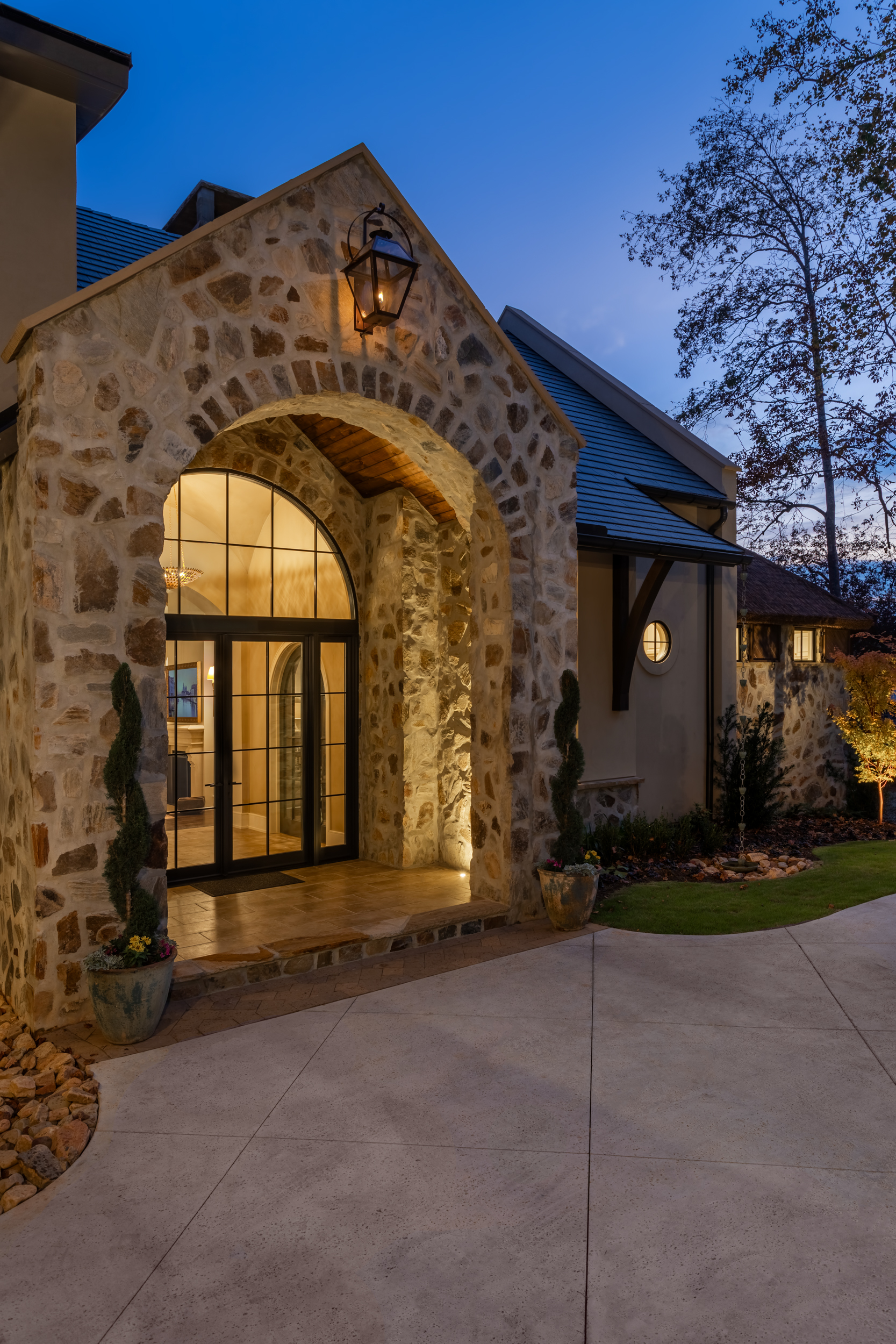 Twilight entry detail at The Pendry Lake Keowee South Carolina &mdash; stone arch entrance with warm interior glow and Endureed Premium Kilimanjaro synthetic thatch turret roof visible in background on luxury lakefront residence