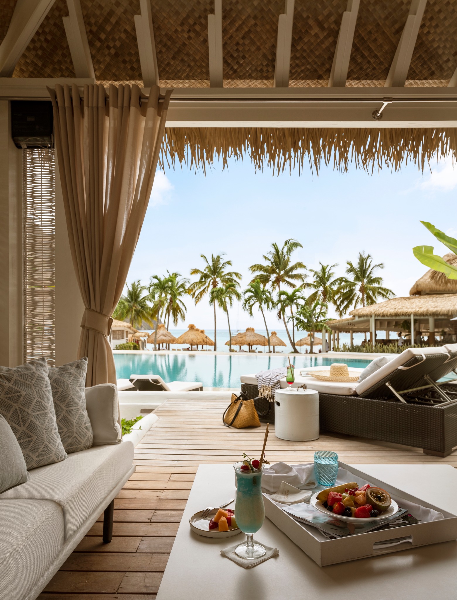 Interior view from Endureed Performance Palm synthetic thatch cabana at Sugar Beach St. Lucia &mdash; engineered thatch eave framing resort pool palm trees beach and Caribbean Sea with styled daybed cocktail and fruit service in foreground