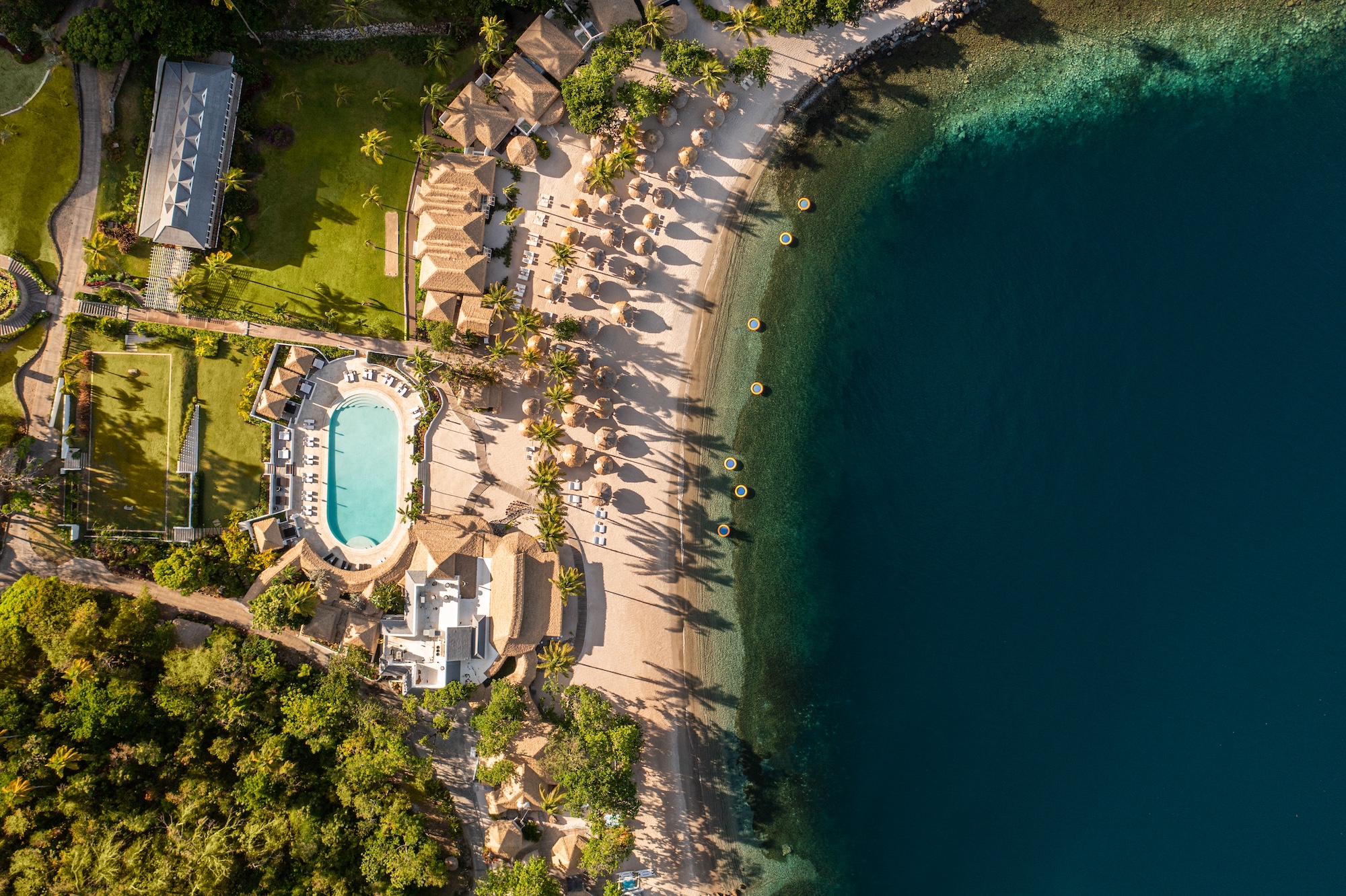 Overhead drone view of Sugar Beach St. Lucia shoreline featuring Endureed Performance Palm synthetic thatch on beach umbrellas cabanas and resort structures &mdash; engineered thatch installation at scale along white sand Caribbean beach with deep blue sea