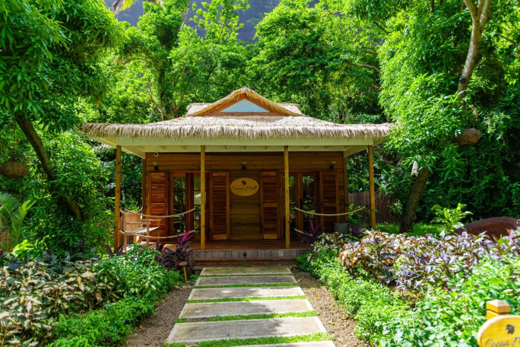 Endureed Performance Palm synthetic thatch on Cocoa Mill building at Sugar Beach St. Lucia &mdash; engineered thatch hip roof with clerestory detail on timber-framed tropical pavilion surrounded by lush resort garden and mountain cliffs