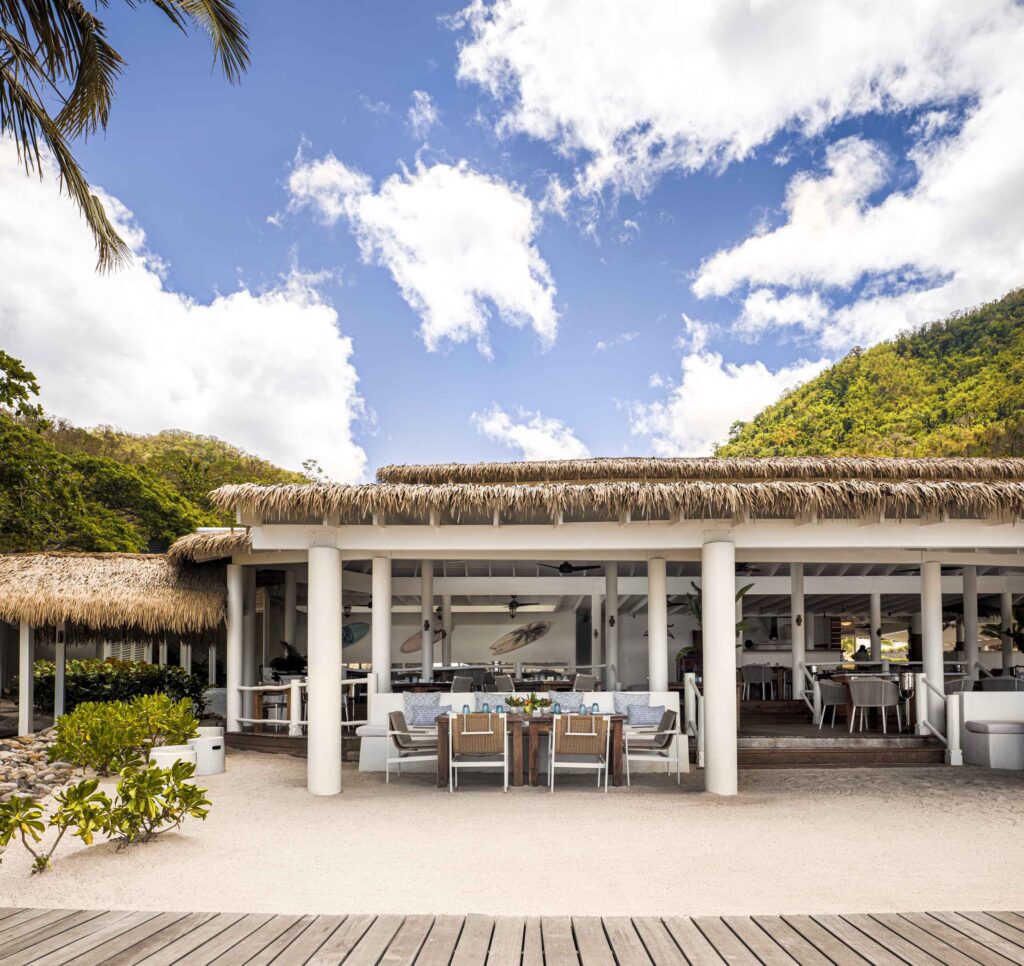 Endureed Performance Palm synthetic thatch on beachfront restaurant and bar at Sugar Beach St. Lucia &mdash; engineered thatch roofline spanning open-air dining pavilion with white columns outdoor dining setup and Caribbean mountain coastline backdrop