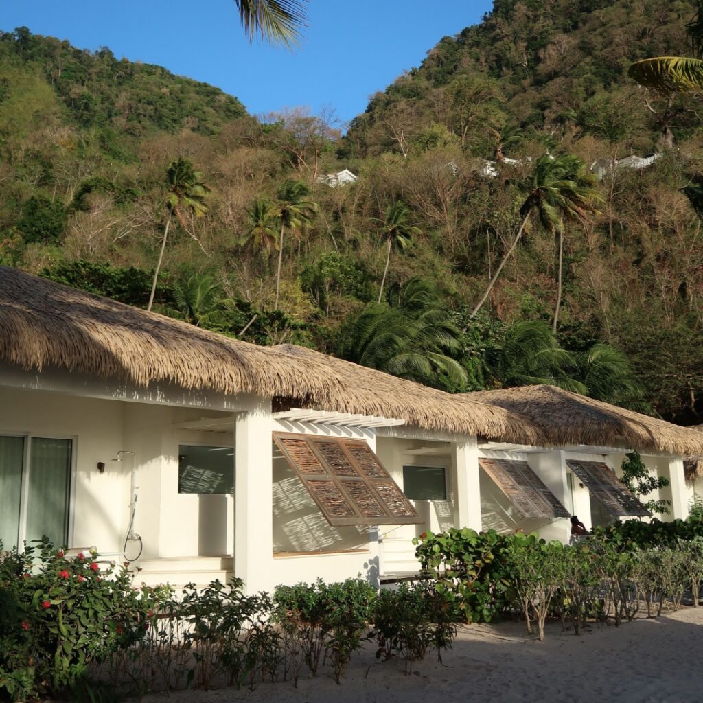 Endureed Performance Palm synthetic thatch roofing on beachfront guest villas at Sugar Beach resort in St. Lucia &mdash; engineered thatch eaves on row of white stucco bungalows with louvered shutters and tropical mountain backdrop