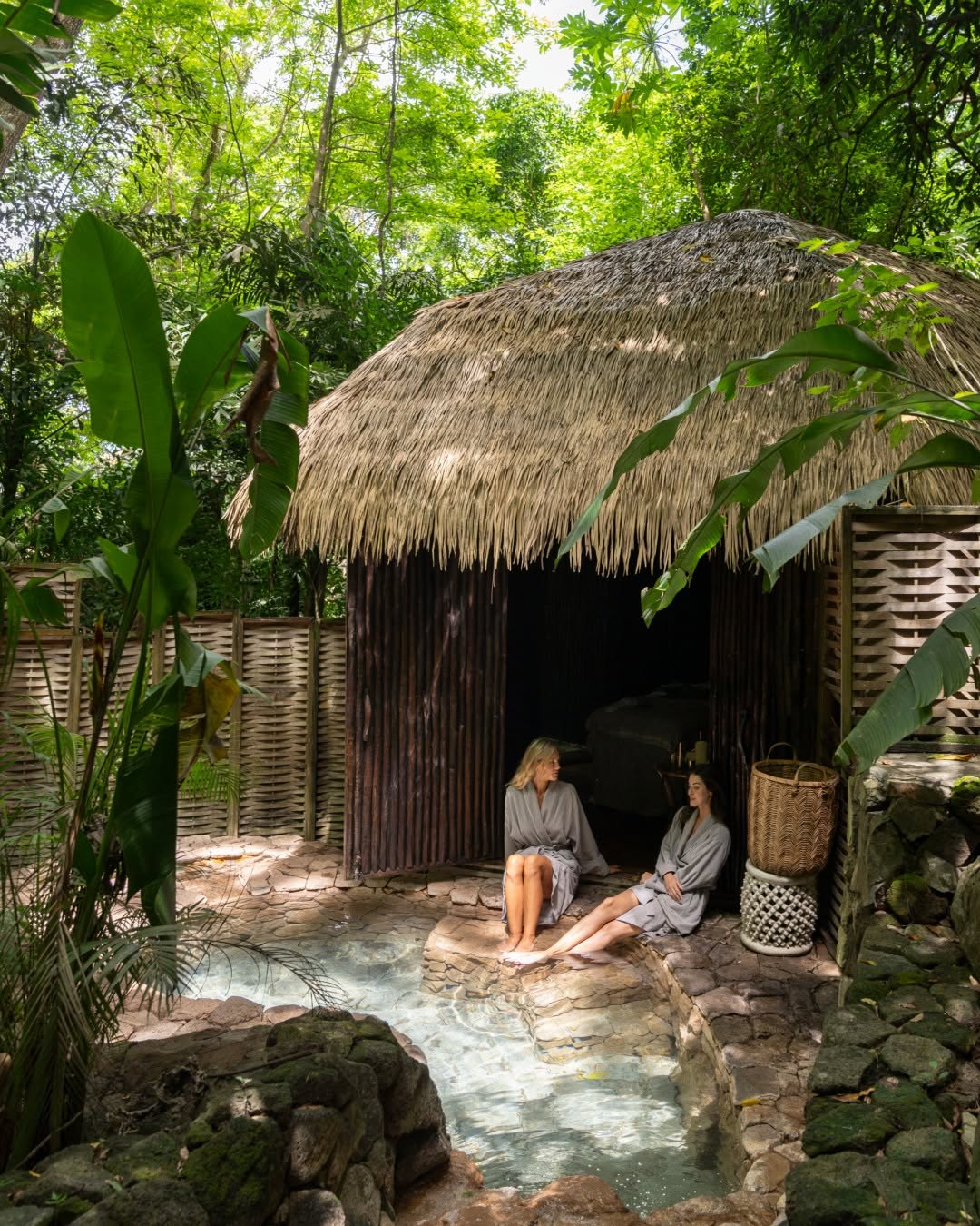 Two guests relaxing at Endureed Performance Palm synthetic thatch spa hut at Sugar Beach St. Lucia &mdash; engineered thatch jungle wellness pavilion with natural stone plunge pool woven bamboo walls and tropical rainforest canopy