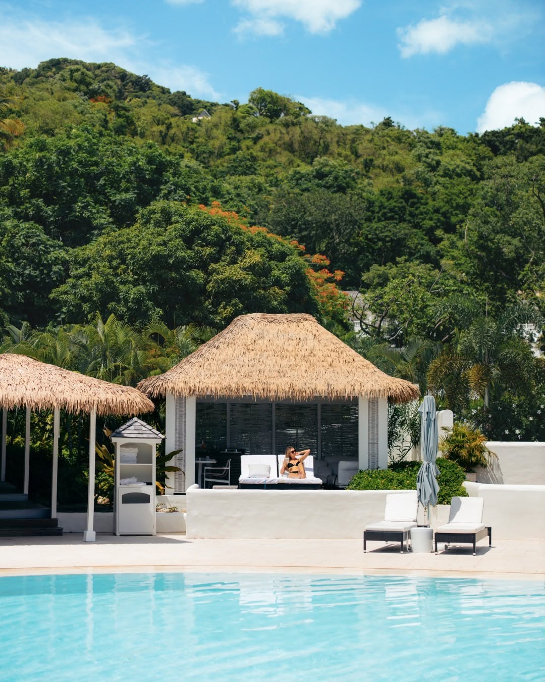 Endureed Performance Palm synthetic thatch pool cabana at Sugar Beach St. Lucia &mdash; guest relaxing under engineered thatch shade structure beside resort pool with tropical hillside and flamboyant tree in background