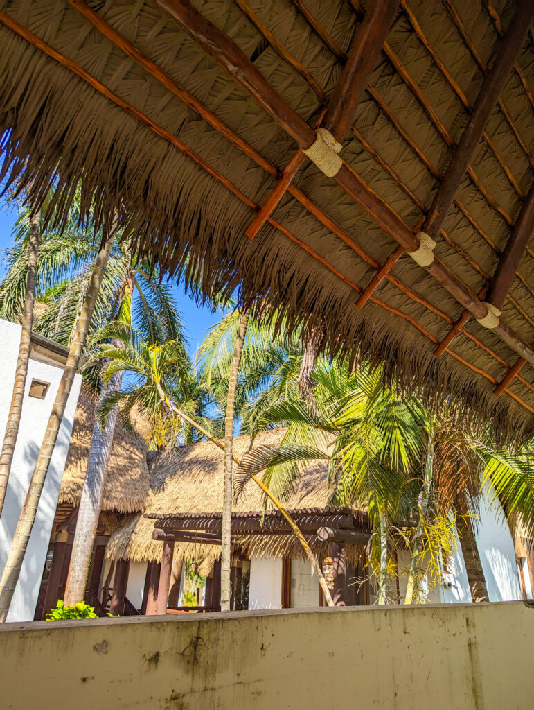 Performance Palm synthetic thatch eave at Casa De Verano, Guatemala, framing a view of palm trees and an adjacent thatched-roof guest casita.