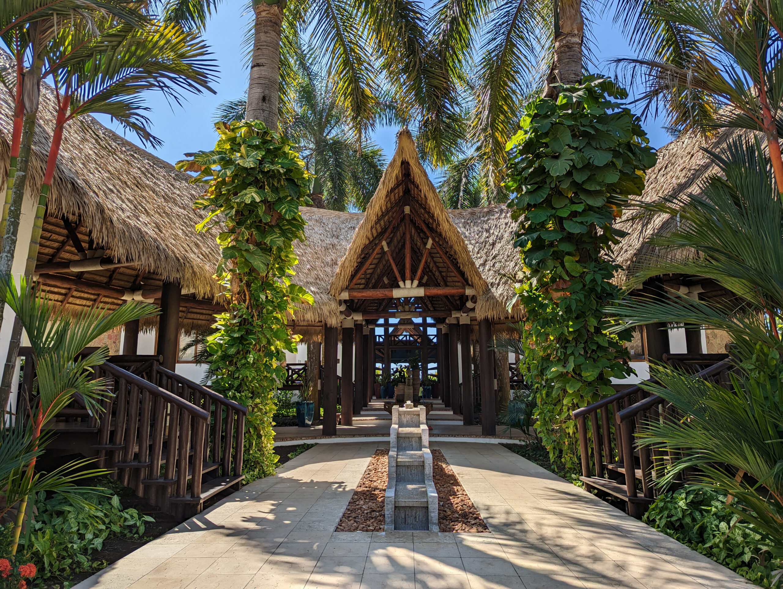 Covered walkway at Casa De Verano leading to a peaked Performance Palm thatch entry pavilion, flanked by tropical landscaping and a center water feature.