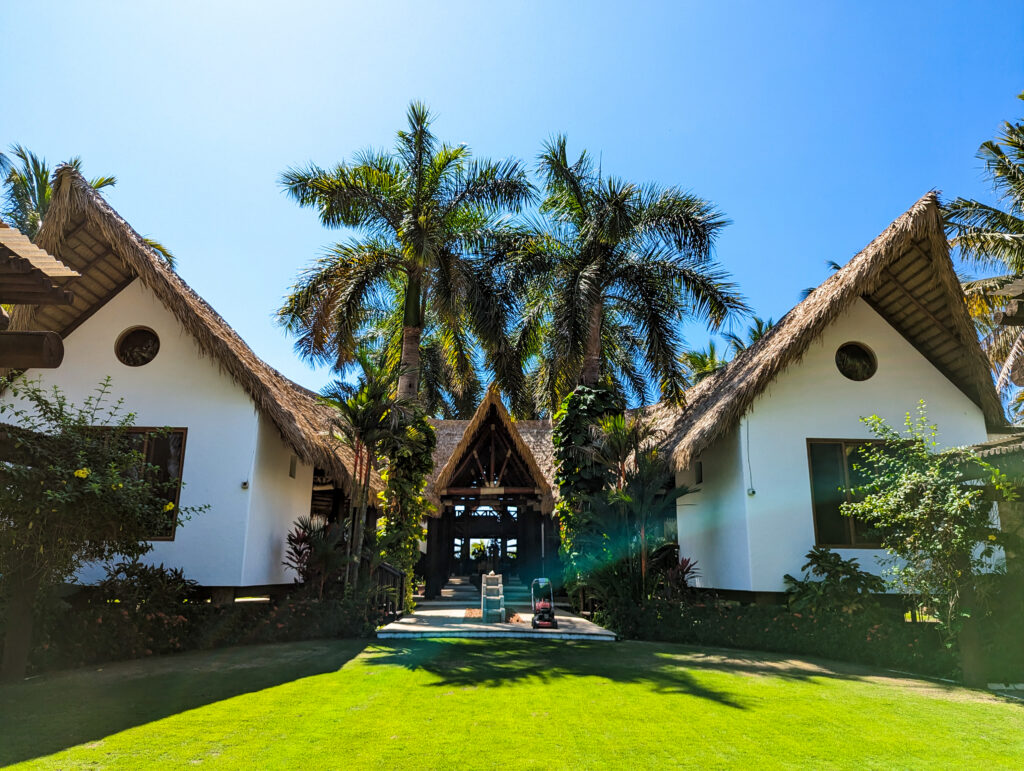 Mirror-image guest casitas at Casa De Verano with Performance Palm thatch gable roofs, connected by a palm-lined lawn and central thatched breezeway.
