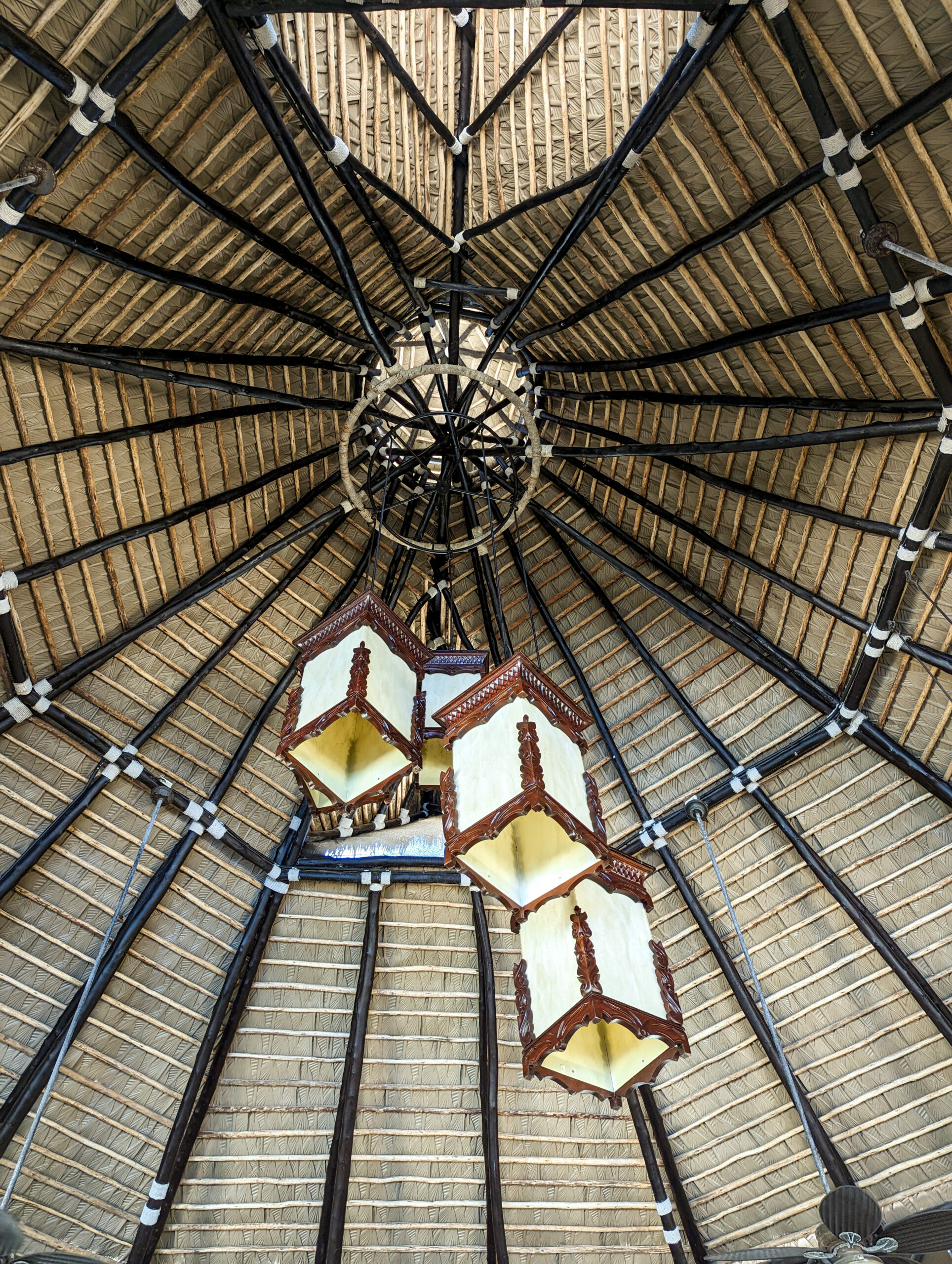 Detailed underside view of a conical Performance Palm thatch roof at Casa De Verano, showing radiating beams and a carved wood lantern fixture at the apex.