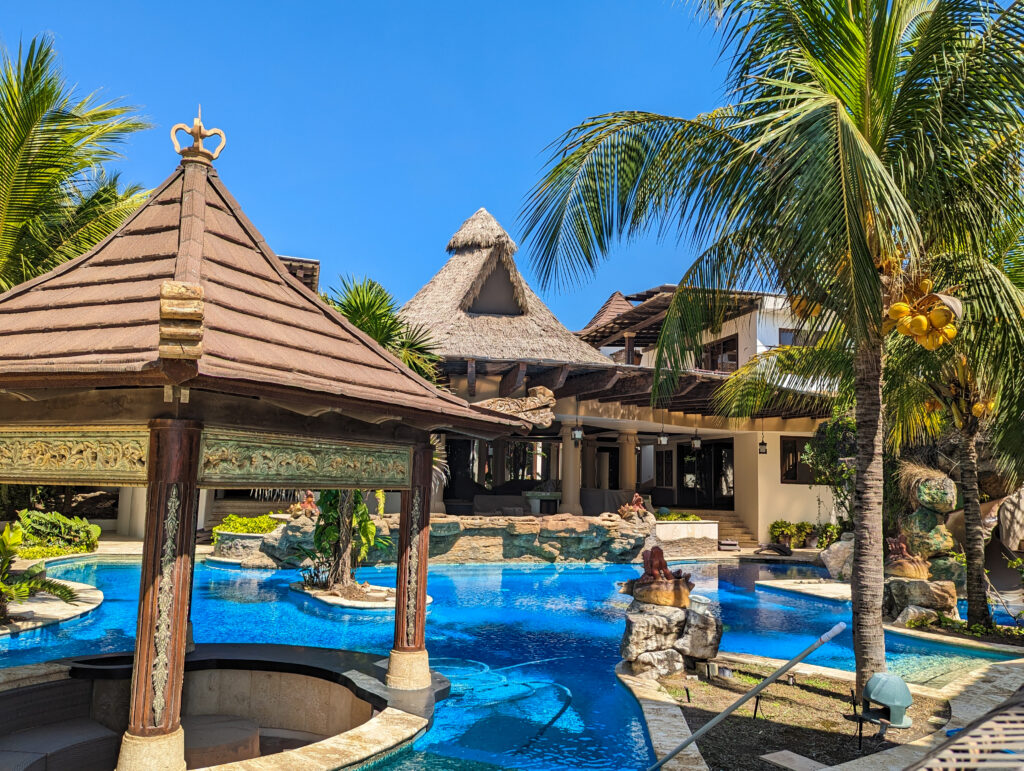 Ornately carved Performance Palm thatch-roofed poolside gazebo at Casa De Verano, with a winding pool, rock waterfall feature, and main house in the background.