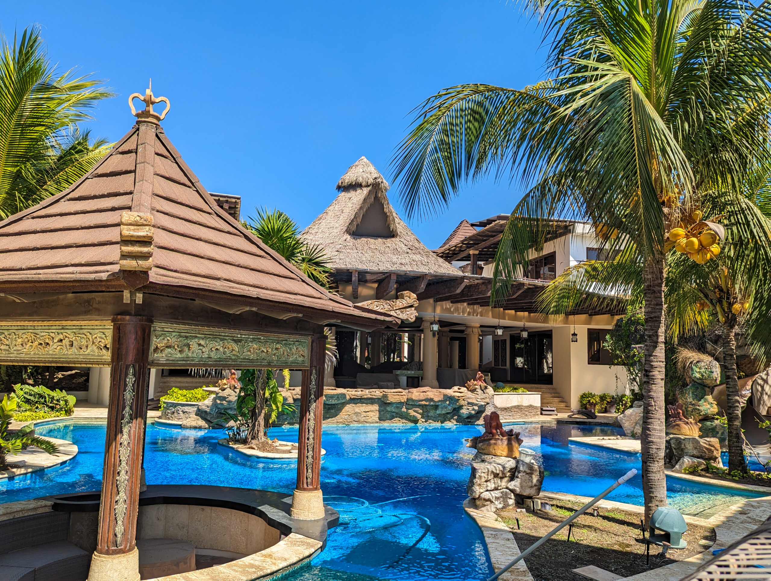 Ornately carved Performance Palm thatch-roofed poolside gazebo at Casa De Verano, with a winding pool, rock waterfall feature, and main house in the background.