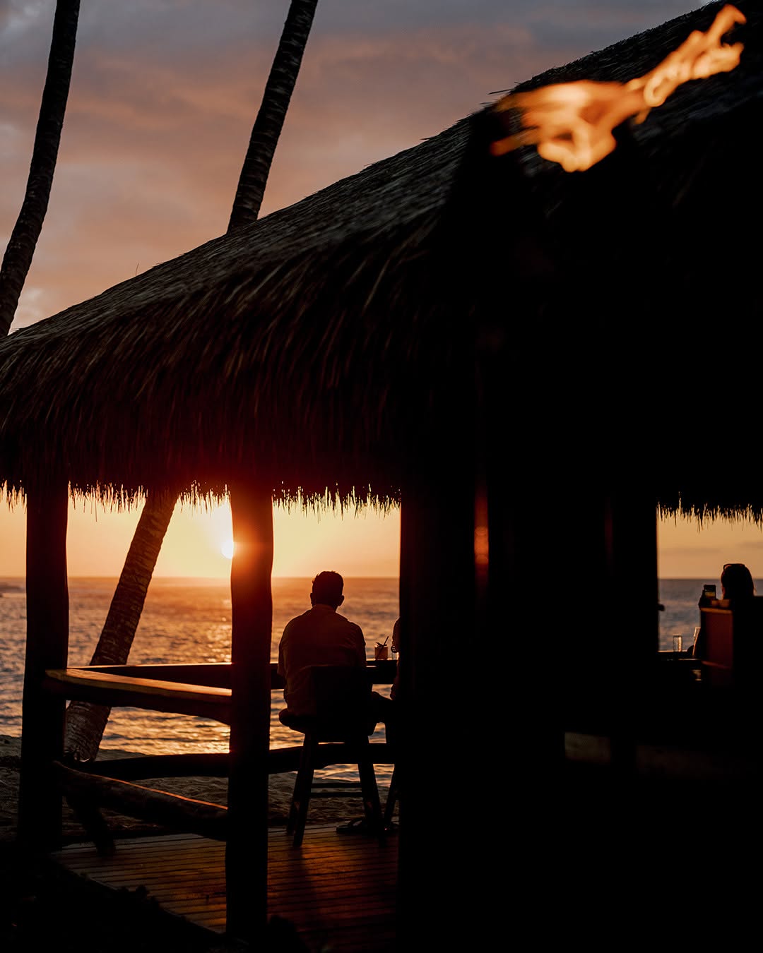 Guests watching the sunset from a thatched-roof bar at Kona Village